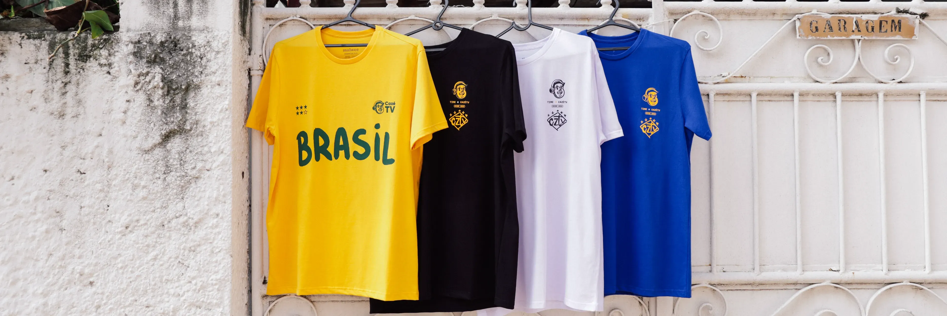 As quatro cores da camisa CazéTV penduradas em cabides à sombra: amarela, preta, branca e azul